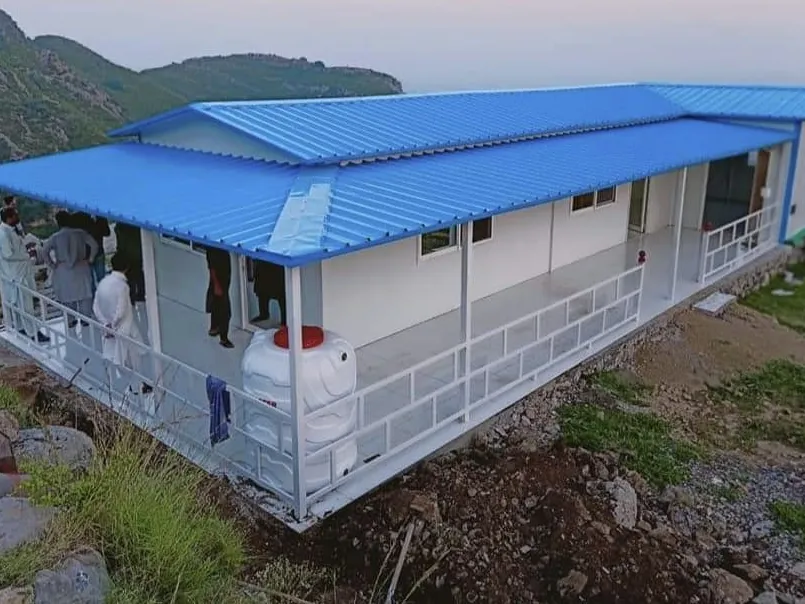 Long white prefabricated building with a blue roof and veranda railing on a hillside