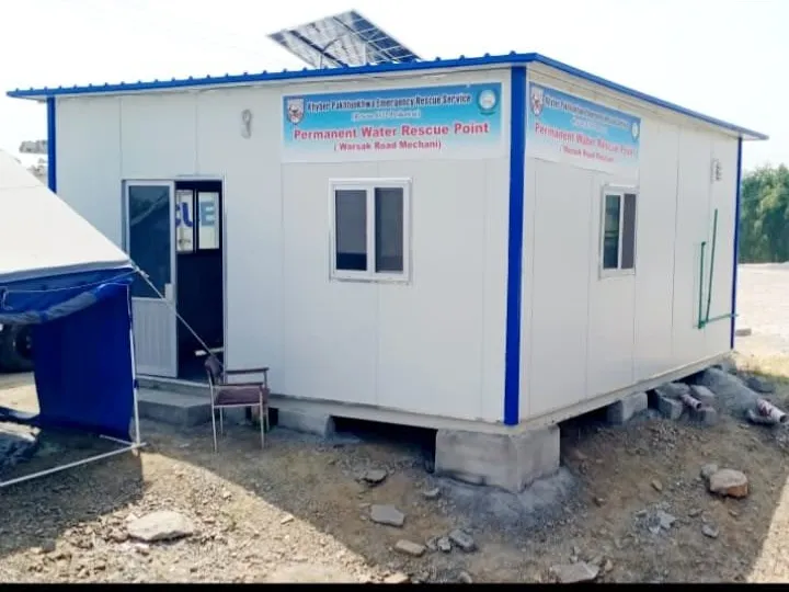 White prefabricated rescue cabin with blue trim, solar panel and front steps on a riverside site