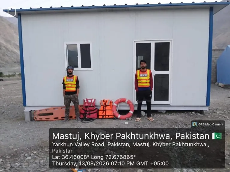 Two rescue workers in high-visibility vests beside a white insulated rescue cabin with rescue equipment, Mastuj